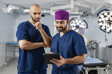 Two surgeons in an operating room review a tablet, likely discussing a patient's medical records before a procedure.
