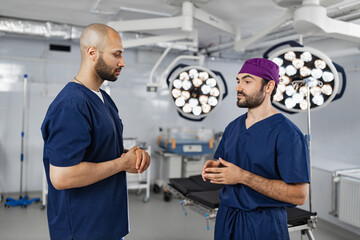Obraz premium Two surgeons in scrubs are engaged in a discussion within a modern operating room, illuminated by surgical lights.