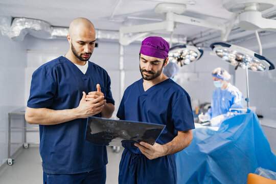 Two surgeons in an operating room examine a medical scan, with another surgeon and medical staff in the background, preparing for a procedure.