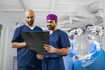 Two surgeons examine a medical scan in an operating room, with other medical staff in the background preparing for a procedure.
