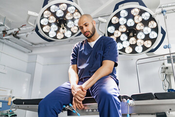 A surgeon sits on an operating table under bright surgical lights, looking thoughtful in a modern operating room.