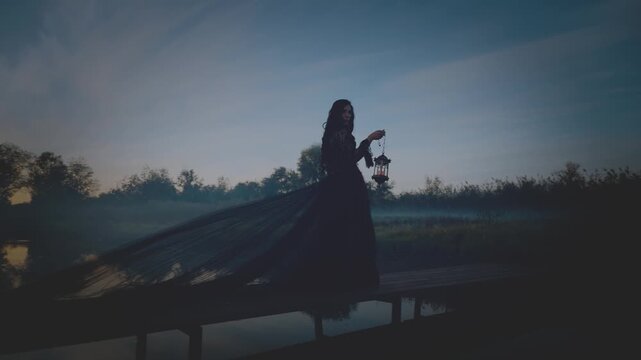 unrecognizable silhouette gothic woman hand hold lantern lamp walking across bridge over river dense fog mist night nature dark dramatic blue sky. long black dress back rear view. Fantasy scared girl