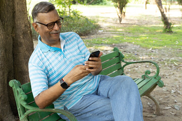 Indian Active happy senior man using and text messaging on mobile while standing at park. Wearing red sweet sweater and eyewear sun glasses. Indian Senior man using digital technology at outdoors