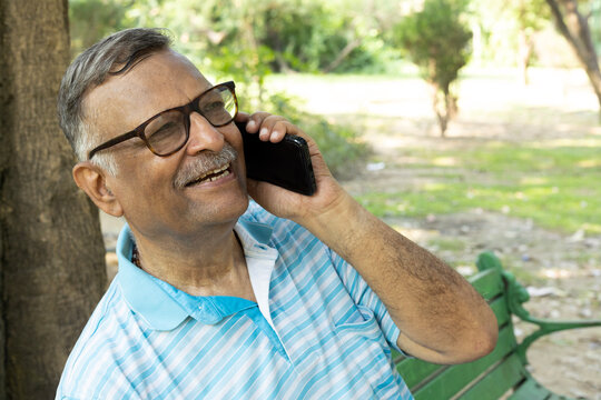 Indian happy senior man talking on mobile at park. Wearing blue t-shirt with eyewear sun glasses. Indian Senior man using digital technology at outdoors