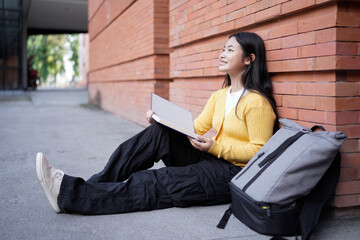 A high school girl sits on the ground her reading a book, surrounded by her backpack and books, reflecting feelings of happy on a university campus