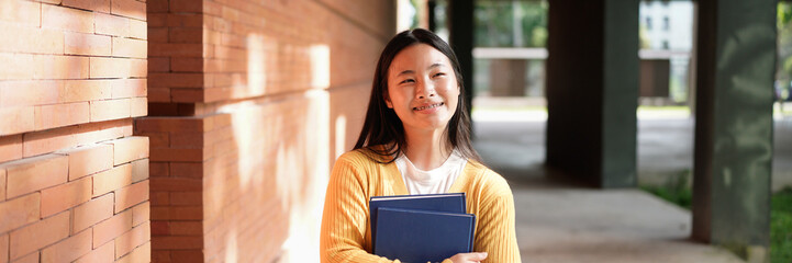 A young woman wearing a yellow sweater and black pants is holding a blue book. She is smiling and she is happy