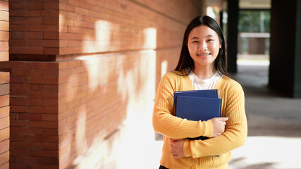 A young woman wearing a yellow sweater and black pants is holding a blue book. She is smiling and she is happy
