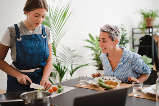Loving mature woman watching young daughter cutting vegetables while spending time and cooking at home