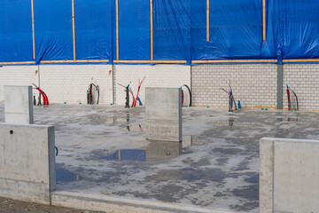 Concrete base plate of a building, with cables and piping coming out of the concrete at the end of the plate. Blue protection plastic sheet above the brick wall.