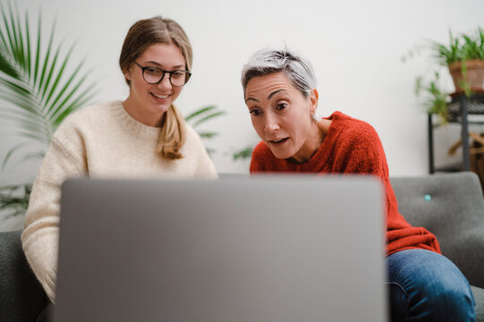 Low angle of excited middle woman looking at screen of laptop while spending time with young daughter at home