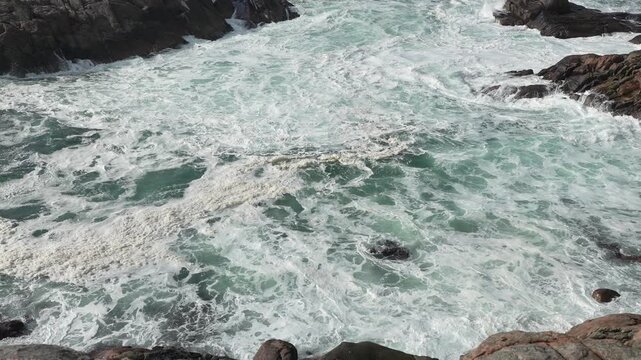 Foamy waves tumble over rugged rocks along the coastline during a bright sunny day. The clear blue water contrasts with dark stones.
