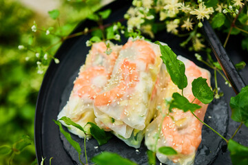 Colorful spring rolls filled with vegetables and shrimp are arranged on a dark, round tray. Lush green plants and delicate flowers surround the dish, creating a lively atmosphere.