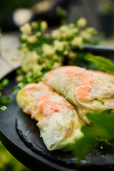 Colorful spring rolls filled with vegetables and shrimp are arranged on a dark, round tray. Lush green plants and delicate flowers surround the dish, creating a lively atmosphere.