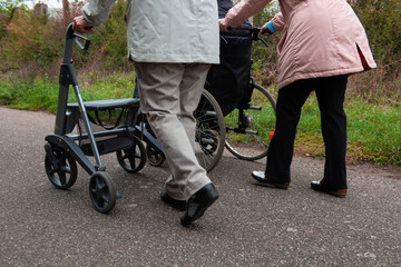Obraz premium Elderly friends taking a walk in the park during cold weather with help of wheelchair and walker enjoying and having fun