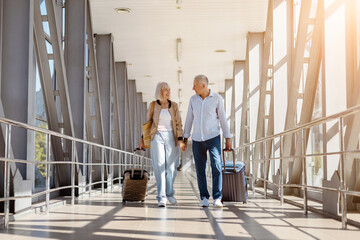 Happy senior couple walking through airport with travel bags, holding hands
