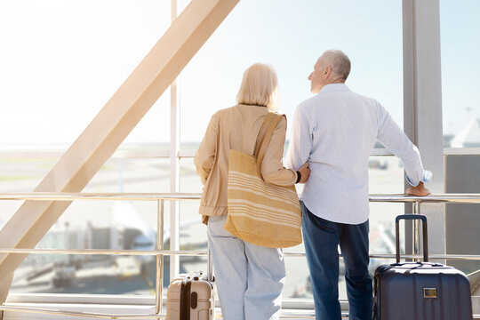 Mature couple standing together looking out of airport window with luggage