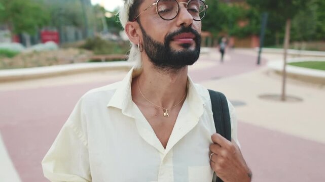 Stylish man with peroxide blonde hair and glasses walks through city. He carries a takeaway coffee cup and a bag over his shoulder. Slow motion.