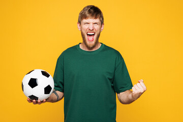 Young happy man fan wear basic green t-shirt cheer up support football sport team holding in hand soccer ball watch tv live stream do winner gesture isolated on plain yellow orange background studio. © ViDi Studio