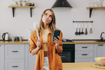 Young happy minded housewife woman wear orange shirt casual clothes use mobile cell phone look for recipe hold wooden spoon sit at table in light kitchen at home alone. Lifestyle cooking food concept.