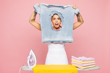Young shocked surprised scared sad woman she wear white apron ironing clothes show damaged t-shirt while doing housework tidy up isolated on plain pastel light pink background. Housekeeping concept.
