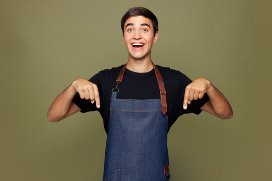 Young man barista bartender barman employee wear jeans apron black t-shirt work in coffee shop point index finger down on area mockup isolated on plain green background Small business startup concept