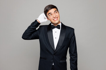 Young sad minded barista male waiter butler man he wears shirt black suit bow tie elegant uniform work at cafe scratch head look aside isolated on plain grey background. Restaurant employee concept.