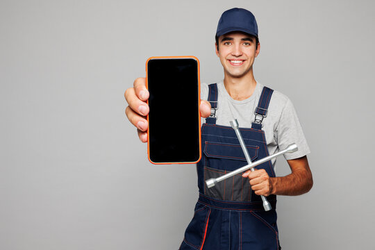 Young car mechanic man wear overall hat uniform workwear clothes work in garage hold crossed wrench tool blank screen mobile cell phone isolated on plain grey background Automotive repair job concept
