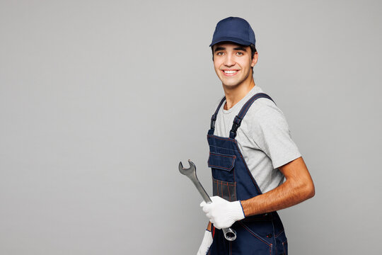 Side view young fun car mechanic man wear overall hat uniform workwear clothes work in garage hold in hand wrench tool looking camera isolated on plain grey background. Automotive repair job concept.