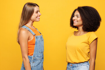 Side profile view two smiling young friends buddies women wear denim casual clothes together meet each other talk speak isolated on plain yellow orange background studio portrait. Lifestyle concept.