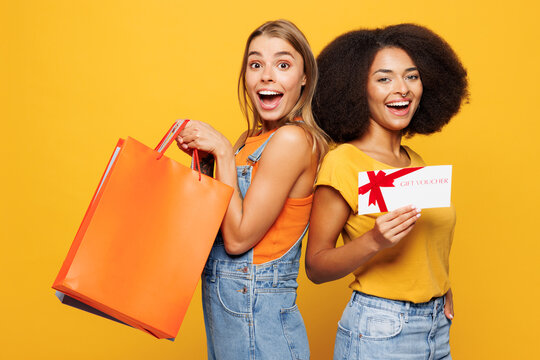 Young two friends women wear casual clothes hold gift coupon voucher card for store shopping paper package bag together isolated on plain yellow orange background. Black Friday sale buy day concept.