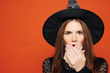 Close up young shocked woman wears black hat dress witch costume cover mouth with hands look camera...