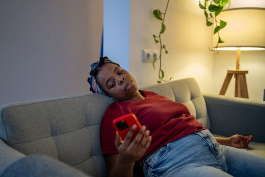 Tired black woman lying on couch with smartphone feeling unmotivated. Weary african american female scrolling social media with bored expression, suffering from digital fatigue, emotional burnout.