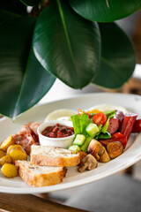 Rustic brunch plate with toast, potatoes, mushrooms, beans, sausage, cheese, and fresh vegetables, styled under green plant leaves in natural light.