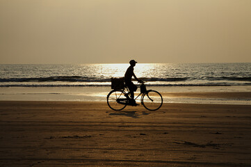 Silhouette of unrecognizable man riding bicycle on seashore, enjoying picturesque view and fresh marine air, on golden beach sand. Water reflecting rays at sunset