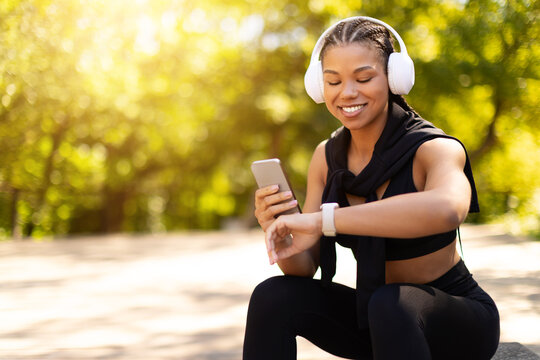 African American woman with headphones sitting outdoors checking smartwatch and holding smartphone