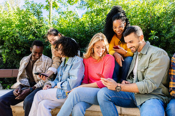 Multiracial young friends using smart phone devices while hanging out at city park. Social media and technology lifestyle concept