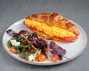 Fresh croissant sandwich with scrambled eggs, melted cheese, and tomato slices served with a colorful side salad of mixed greens, feta, and cherry tomatoes