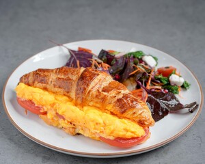 Fresh croissant sandwich with scrambled eggs, melted cheese, and tomato slices served with a colorful side salad of mixed greens, feta, and cherry tomatoes