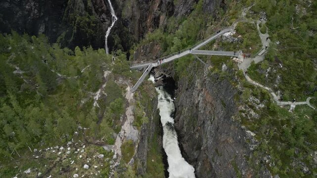 Drone shot of the bridge at the tourist attraction V&oslash;ringsfossen Waterfall in Norway. People walk across the bridge and cross a steep valley through which the water flows to the waterfall.