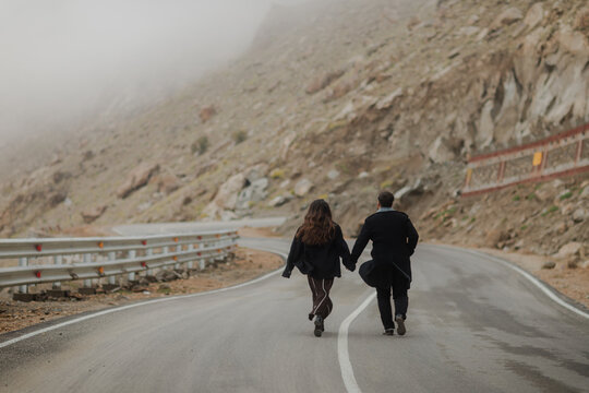 Couple holding hands runs down winding mountain road, back view, hazy mountain scenery in background, symbolizing adventure and togetherness.