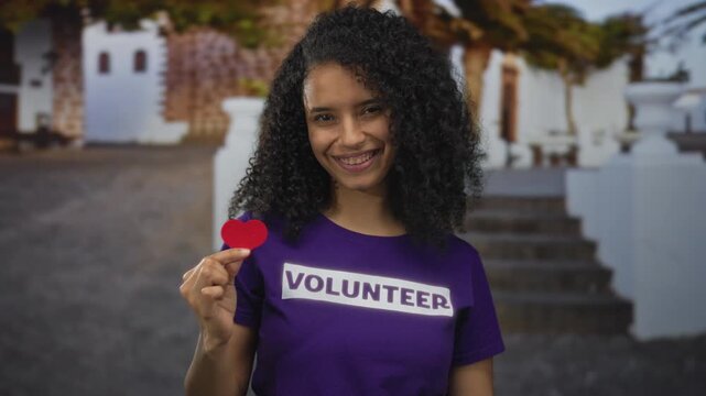Young woman holding paper heart wearing volunteer shirt outdoors with a smile showcasing community spirit and kindness in a vibrant urban setting with colonial architecture.