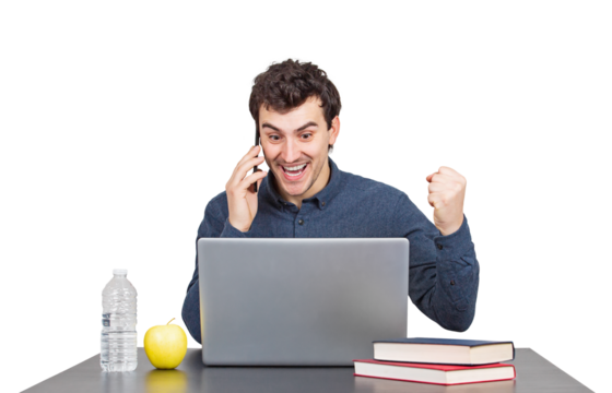 Excited young man seated at the working desk looking contented in his laptop, talking on phone and raises up his fist, achievement and celebration gesture. Successful male isolated portrait