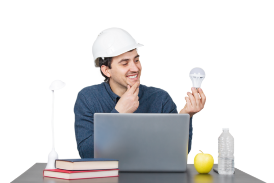 Contented young man engineer wearing white helmet, sitting at the working desk looks thoughtful at a lightbulb in his hand, isolated on transparent background. Engineering idea concept