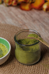 A wooden spoon holds a scoop of bright green matcha powder over a glass container filled with more powder.