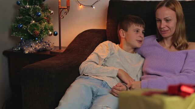 Loving mom and teenage son relaxing on sofa, chatting and sharing smiles in their festive living room adorned with tiny christmas tree and twinkling lights during holiday season