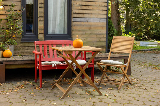 Wooden table with pumpkin, red bench and chair on backyard patio in daylight.