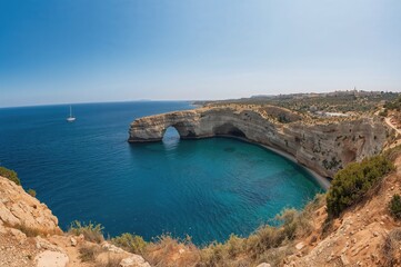 Wide-angle view of a coastal bay featuring a notable rock formation, clear blue waters, and a sailboat under a bright sunny sky