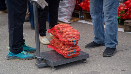 Fresh farm carrots in red mesh sacks sit on a platform scale at an outdoor farmers market; buyers’ and seller’s feet frame the street‑trade weighing scene. Photo