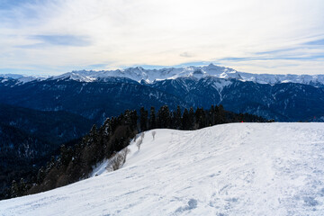 Panoramic view of the highlands, valley, forest and snowy slopes. Mountains covered with snow are visible in the distance. The atmosphere of a winter resort.