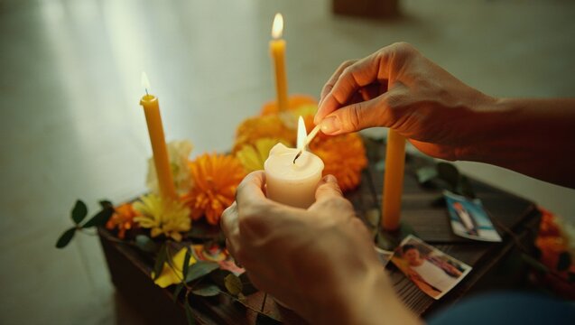 Hands lighting a white candle on an altar of orange marigolds and framed photos, a solemn remembrance ritual honoring deceased loved ones with quiet devotion and hope